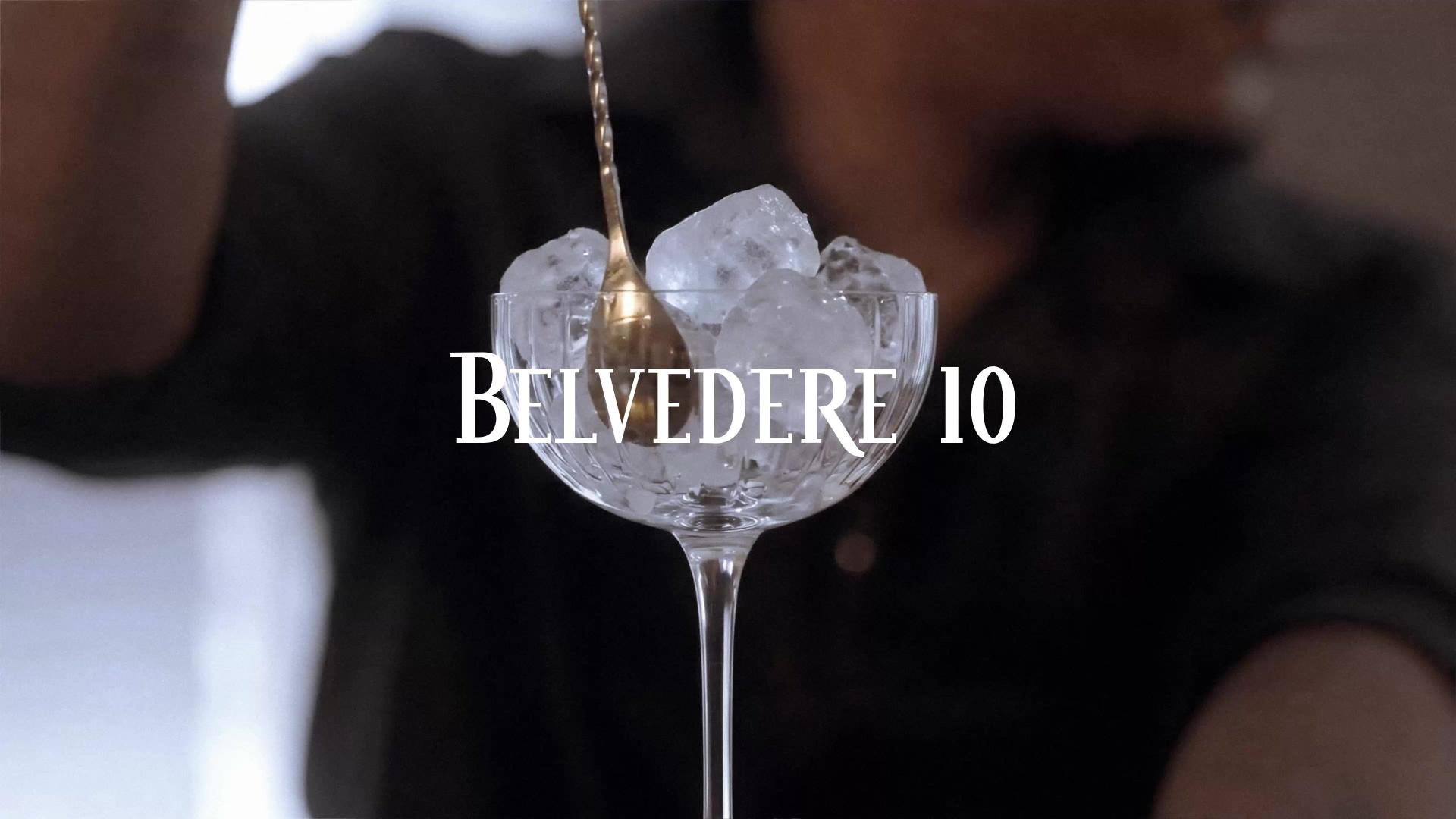 A bar spoon lowered into a crystal coupe filled with cracked ice, bartender at work behind