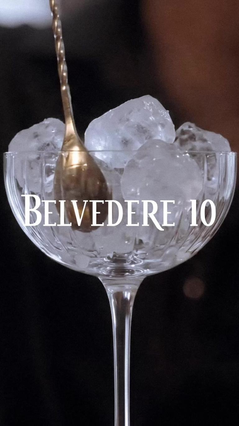 A bar spoon lowered into a crystal coupe filled with cracked ice, bartender at work behind